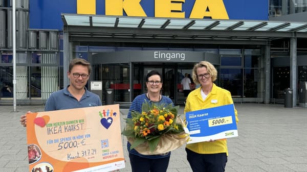 Drei Frauen stehen vor dem IKEA-Gebäude in Kaarst und halten gemeinsam einen Spendenscheck über 5.000 Euro. Die mittlere Frau trägt eine gelbe IKEA-Uniform.