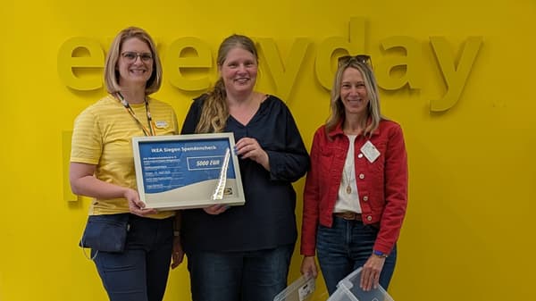 Drei Frauen stehen vor einer gelben Wand mit dem Schriftzug „everyday“. Die Frau links trägt ein gelbes IKEA-Shirt mit Namensschild, zusammen mit der Frau in der Mitte hält sie einen großen gerahmten Spendenscheck über 5.000 Euro von IKEA Siegen für den Kinderschutzbund. Die Frau rechts trägt eine rote Jacke und hält zwei transparente Aufbewahrungsboxen in der Hand.