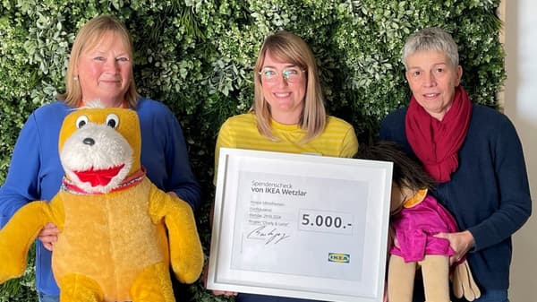 Drei Frauen stehen vor einer grünen Pflanzenwand und halten einen großen Spendenscheck von IKEA Wetzlar über 5.000 Euro. Die Frau in der Mitte trägt ein gelb-gestreiftes Oberteil und hält den Scheck, während die beiden anderen Frauen je eine große Stoffpuppe in den Händen halten.  