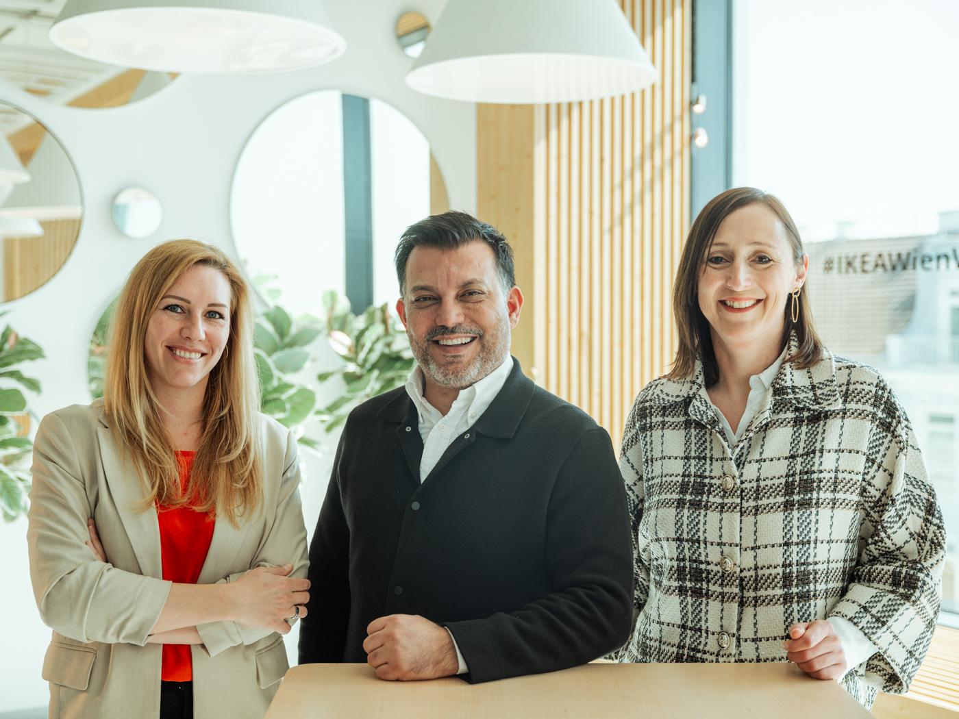 Drei Führungskräfte von IKEA stehen in einem modernen Besprechungsraum mit Blick über die Dächer einer Stadt – professionelles Team im urbanen Arbeitsumfeld.

