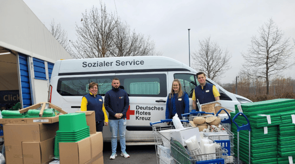 Drei IKEA-Mitarbeitende in blauer und gelber Kleidung sowie ein Vertreter des Deutschen Roten Kreuzes stehen vor einem weißen DRK-Transporter mit der Aufschrift „Sozialer Service“. Vor ihnen sind zahlreiche Kartons, Einkaufswagen mit Haushaltsartikeln und gestapelte grüne Matten zu sehen.  