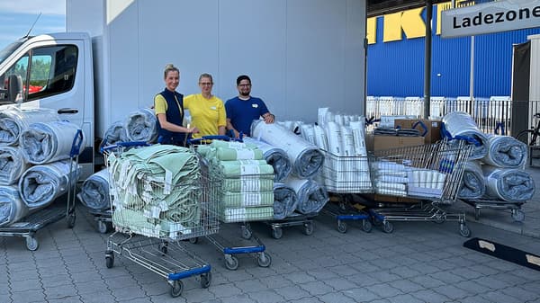 Drei IKEA-Mitarbeitende stehen vor der Ladezone eines IKEA-Gebäudes neben einem weißen Transporter. Sie sind umgeben von Einkaufswagen voller gespendeter Bettwaren, darunter Decken, Matratzenauflagen und Handtücher in Grün und Grau. Im Hintergrund ist die blaue Fassade mit dem gelben IKEA-Logo zu sehen.  