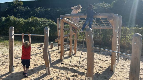Drei Kinder spielen auf einem Spielplatz mit Klettergerüst aus Holz und einer Reckstange. Der Boden ist mit Sand bedeckt, im Hintergrund sind grüne Büsche und ein hoher Zaun zu sehen.