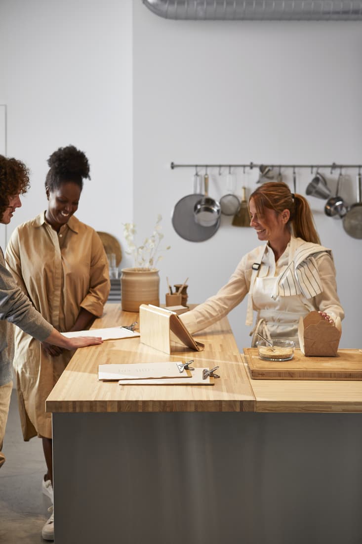 Drei Personen an Küchentheke mit Utensilien im Hintergrund, eine Person mit Schürze und Papiertüte.