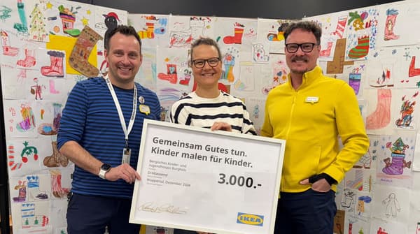 Drei Personen halten einen grossen IKEA-Spendenscheck in Höhe von 3'000 Euro mit der Aufschrift „Gemeinsam Gutes tun. Kinder malen für Kinder.“ Hinter ihnen ist eine Wand mit vielen bunten Kinderzeichnungen zu sehen. Zwei der Personen tragen IKEA-Dienstkleidung, eine in Blau, die andere in Gelb. Die dritte Person trägt einen gestreiften Pullover. Alle lächeln in die Kamera.  