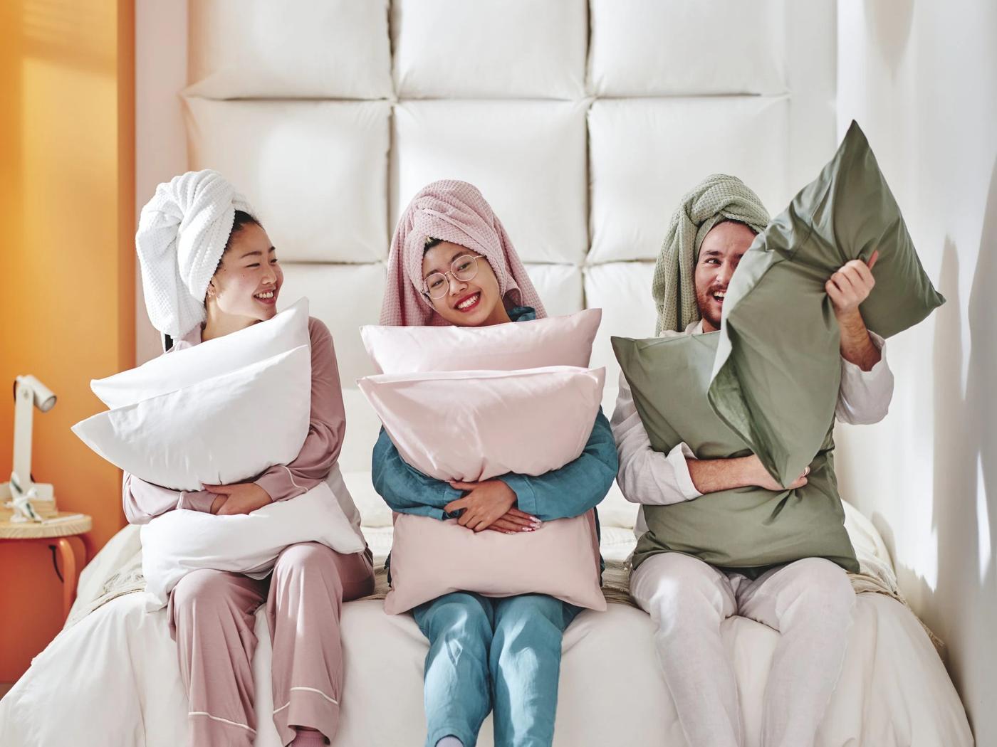 Drei Personen in Schlafanzügen und mit Handtüchern um den Kopf sitzen lachend auf einem Bett und kuscheln mit farbigen IKEA-Kissen – fröhliche Morgenstimmung mit Fokus auf Gemütlichkeit und Wohlfühltextilien.