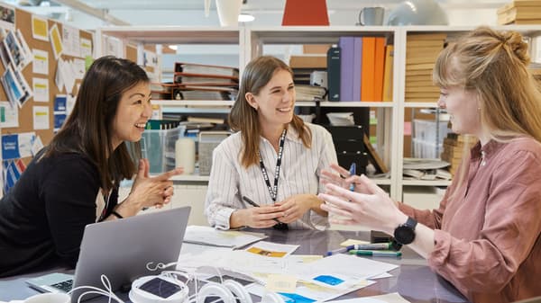 Drei Personen sitzen an einem Schreitisch und unterhalten sich. Auf dem Schreitisch liegt Papier, Muster und ein Laptop.