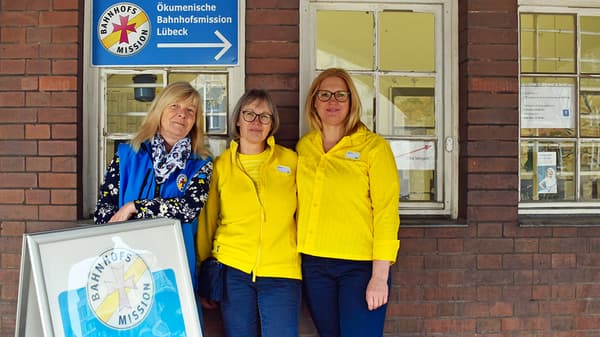 Drei Personen stehen dicht beieinander vor einem Backsteingebäude mit zwei alten, weißen Fensterrahmen. Links im Bild befindet sich ein Schild der Bahnhofsmission Lübeck sowie ein Aufsteller mit dem Logo der Bahnhofsmission. Zwei der Personen tragen gelbe Oberteile, eine Person trägt eine blaue Weste über einem gemusterten Oberteil. Alle stehen frontal zur Kamera.