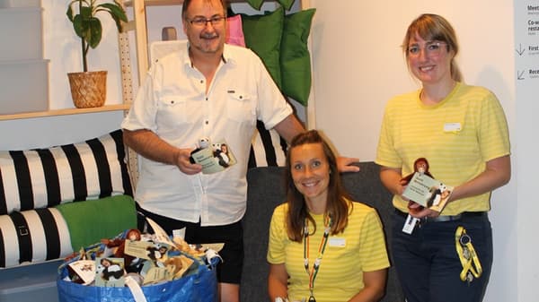 Drei Personen stehen in einem IKEA-Einrichtungshaus und präsentieren kleine Stofftiere mit Spendenanhängern. Zwei von ihnen tragen gelbe IKEA-Shirts, während der Mann in einem weissen Hemd eine Spendenaktion unterstützt. Im Vordergrund steht eine grosse blaue IKEA-Tasche voller Stofftiere.