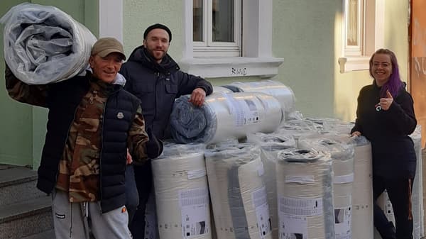 Drei Personen stehen vor einem grünen Gebäude mit einer großen Anzahl an aufgerollten, verpackten Matratzen von IKEA. Eine Person trägt eine auf der Schulter, während eine andere den Daumen hochhält. Alle lächeln.