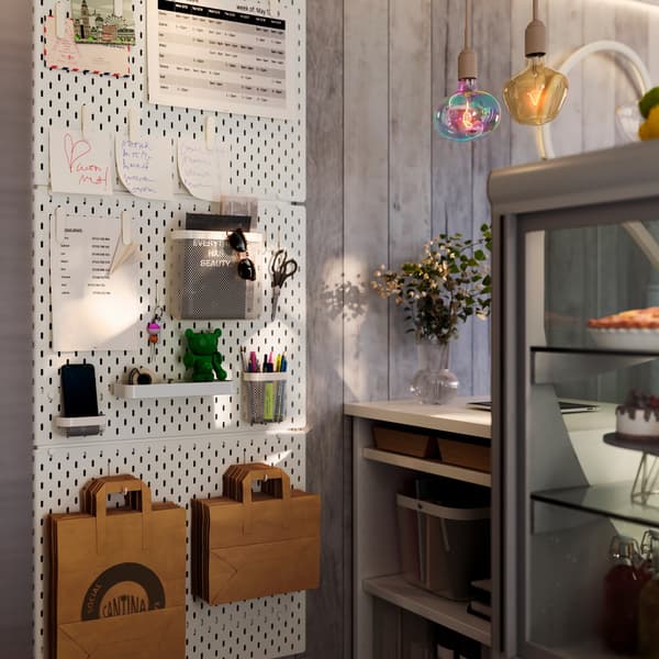 Drei SKÅDIS Lochplatten sind an der Wand neben der Theke eines Cafés montiert. An Haken hängen Einkaufstaschen.
