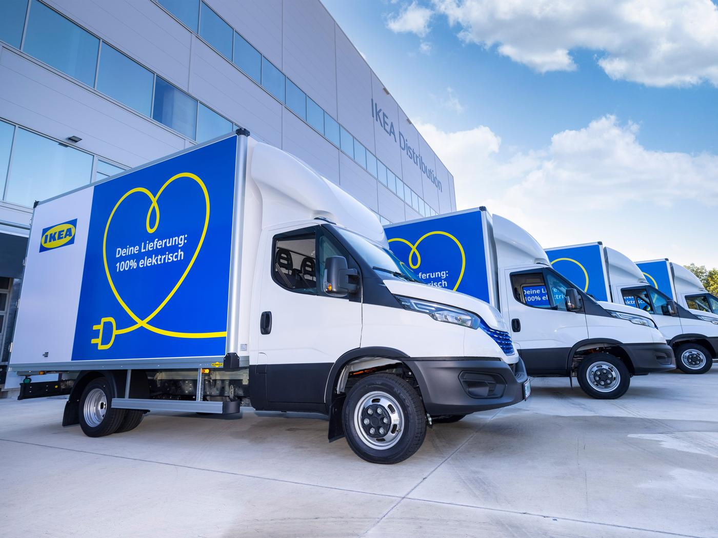 Drei weiße Elektro-Lieferwagen von IKEA parken vor einem IKEA Distributionszentrum unter einem blauen Himmel. Die Lieferwagen sind blau und weiß lackiert mit einem gelben Herz-Logo, das einen Stecker umgibt, und der Aufschrift "Deine Lieferung: 100% elektrisch"
