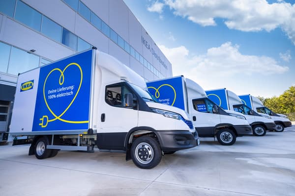 Drei weiße Elektro-Lieferwagen von IKEA parken vor einem IKEA Distributionszentrum unter einem blauen Himmel. Die Lieferwagen sind blau und weiß lackiert mit einem gelben Herz-Logo, das einen Stecker umgibt, und der Aufschrift "Deine Lieferung: 100% elektrisch"