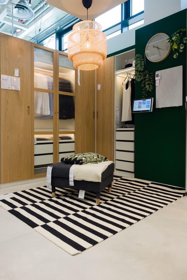 Dressing modern cu uși din lemn și oglinzi, covor alb-negru în dungi, taburet gri cu pătură verde și albă, lampă din ratan, perete verde cu oglindă rotundă și plante decorative.