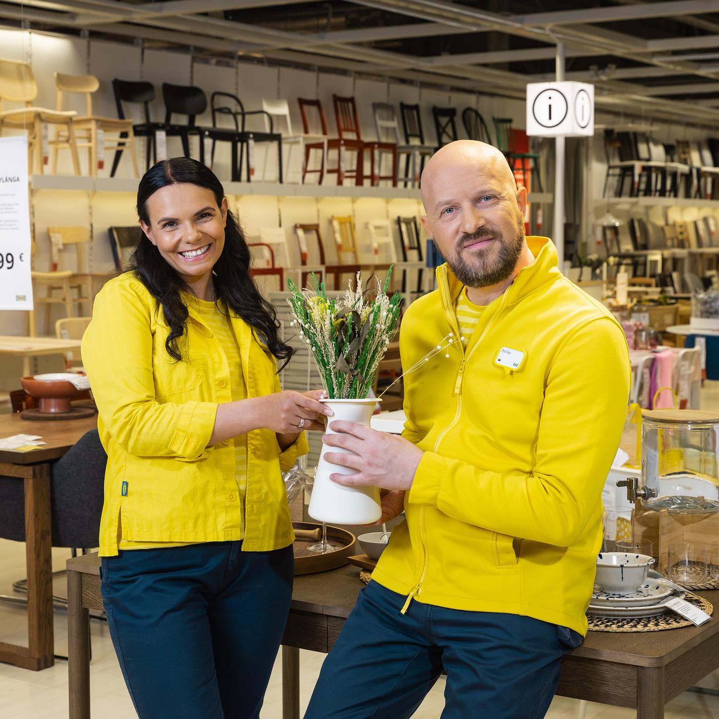 Du IKEA ekspertai su geltonos ir mėlynos spalvų uniformomis stovi Valgomojo baldų skyriuje ir kartu laiko baltą vazą su gėlėmis.
