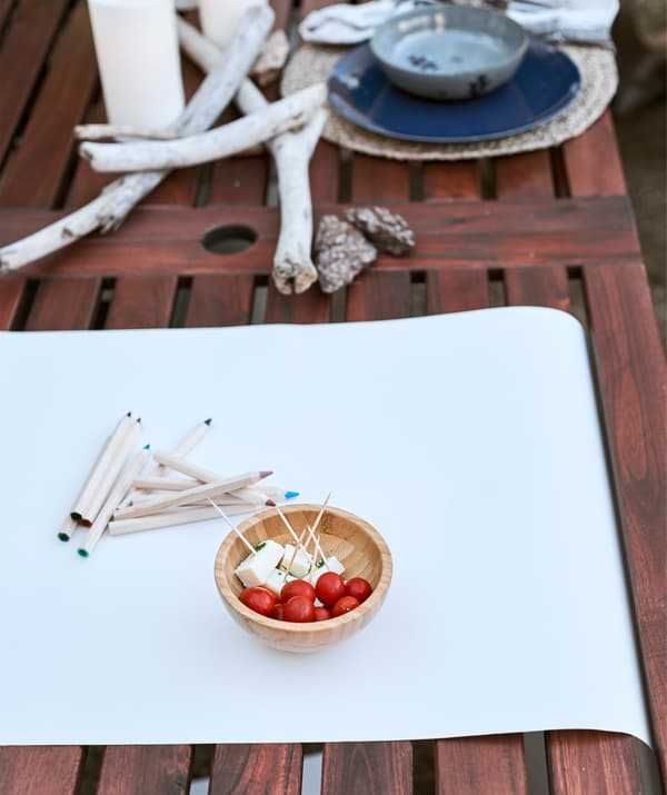 Du papier blanc et des crayons de couleur et un petit bol de choses à grignoter sur une table en bois, du bois flotté et un set avec couvert.