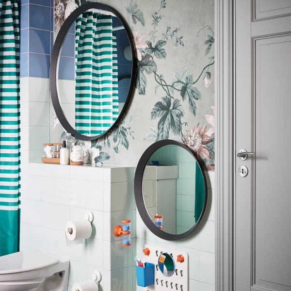 Dua cermin bulat dengan saiz yang berbeza tergantung pada ketinggian yang berbeza pada dinding yang mempunyai kertas dinding berbunga-bunga di dalam bilik mandi.