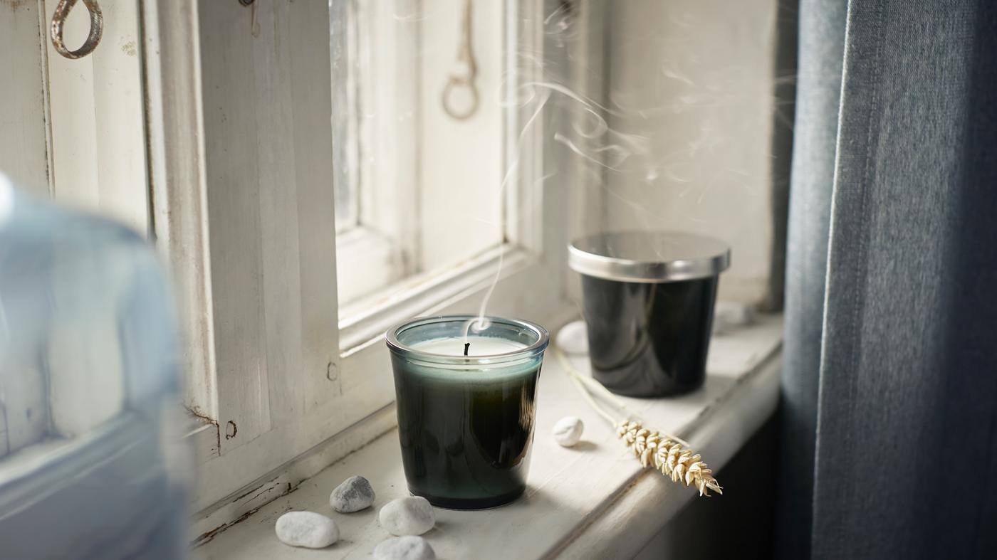 Dua lilin wangi di dalam gelas PÄRONTRÄD berwarna firus terletak di ambang tingkap dengan batu kerikil berwarna putih bertaburan di atasnya.