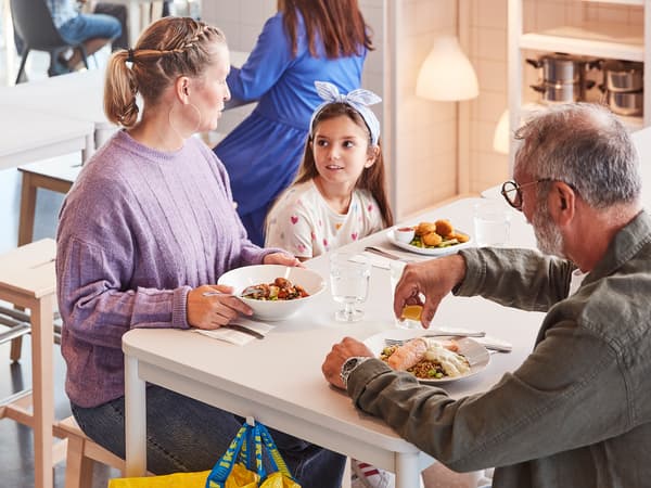 Dua orang dewasa dan kanak-kanak sedang menikmati hidangan bersama-sama sambil duduk di meja berwarna putih di Bistro Sweden IKEA.
