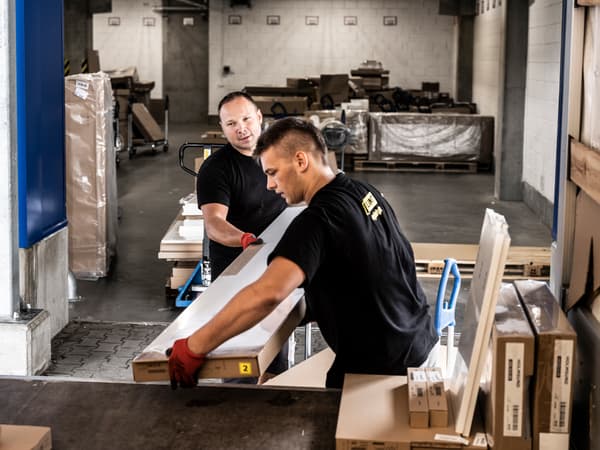 Dua orang lelaki yang memakai kemeja-T berwarna hitam dan sarung tangan kerja berwarna merah sedang memunggah pek leper IKEA dari troli ke dalam bahagian belakang trak.