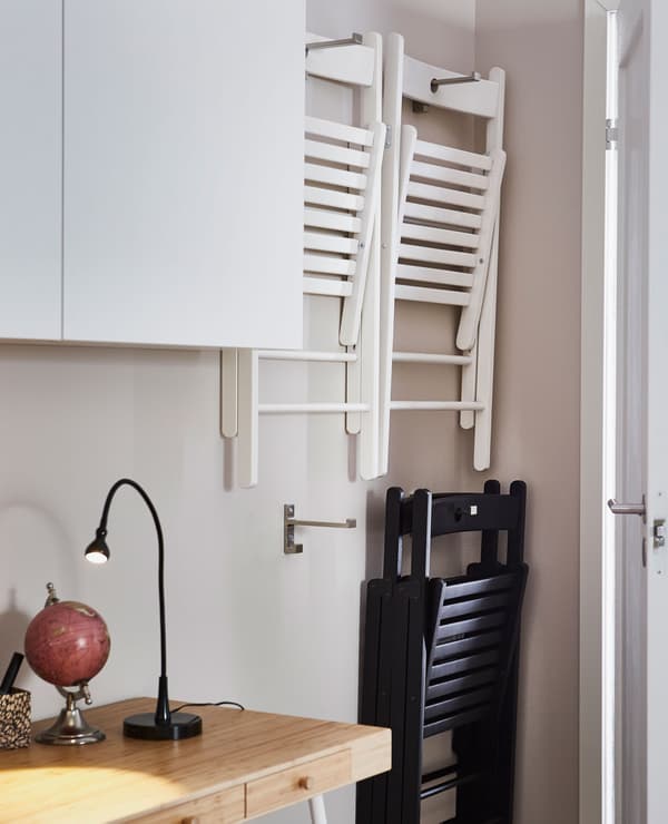 Dúas cadeiras pregables colgadas da parede en ganchos de aceiro inoxidable BJÄRNUM tras unha porta do salón.