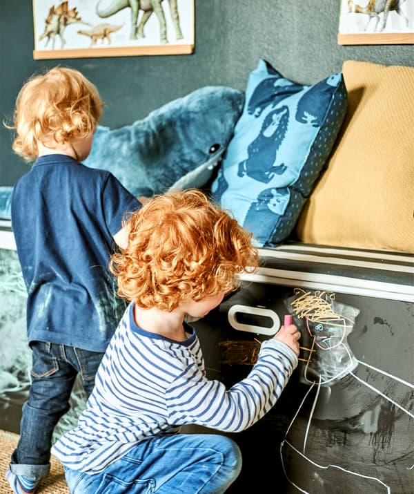 Due bambini disegnano con un gessetto su una cassettiera dipinta con vernice effetto lavagna.