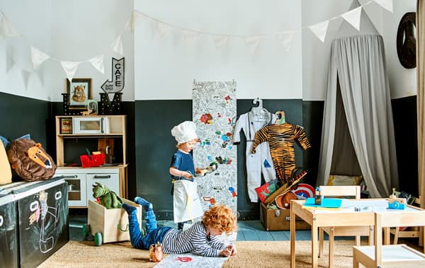 Due bambini giocano in una stanza con un tavolo e sedie per bambini LÄTT, un contenitori per giocattoli FLISAT, una cucina gioco DUKTIG e bandierine decorative - IKEA