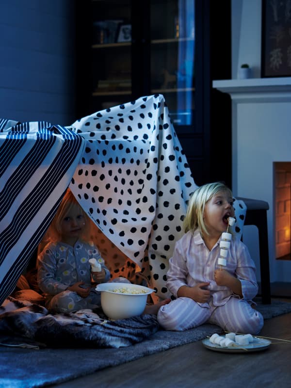 Due bambini in pigiama seduti sul pavimento mentre stanno mangiando dei marshmallows e pop corn. Accanto, tessuti che ricreano delle tende.