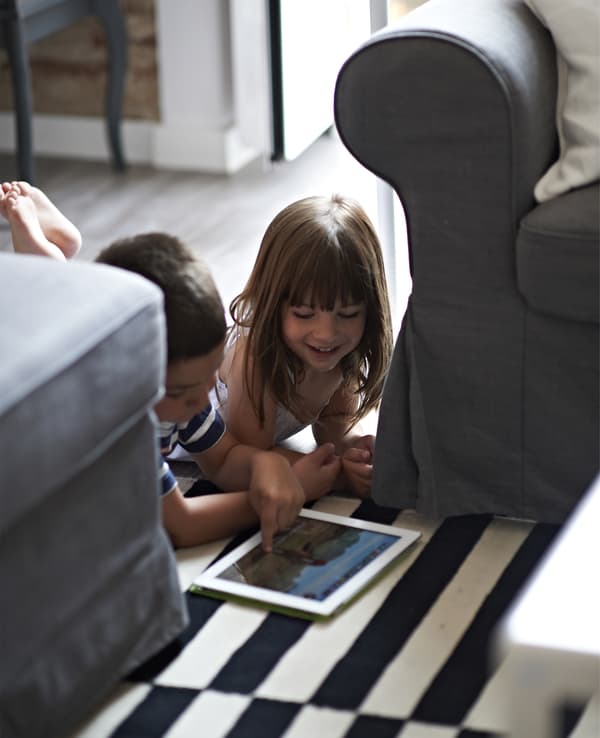 Due bambini sdraiati sul pavimento tra due divani che giocano con un tablet.