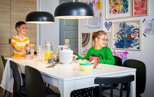 Due bambini seduti a un tavolo bianco, due lampade a sospensione nere e una collezione di disegni incorniciati alla parete alle loro spalle.