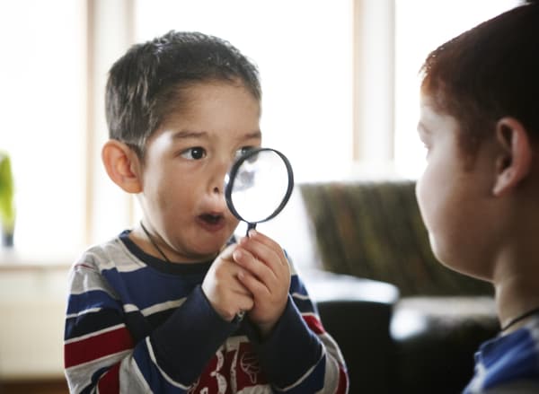 Due bambini seduti l’uno di fronte all’altro, uno dei quali fa delle facce divertenti guardando attraverso una lente di ingrandimento.
