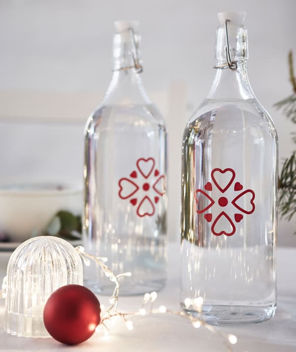 Due bottiglie di vetro trasparente con piccoli decori rossi su un tavolo accanto a una luce decorativa a LED e fili di luci - IKEA