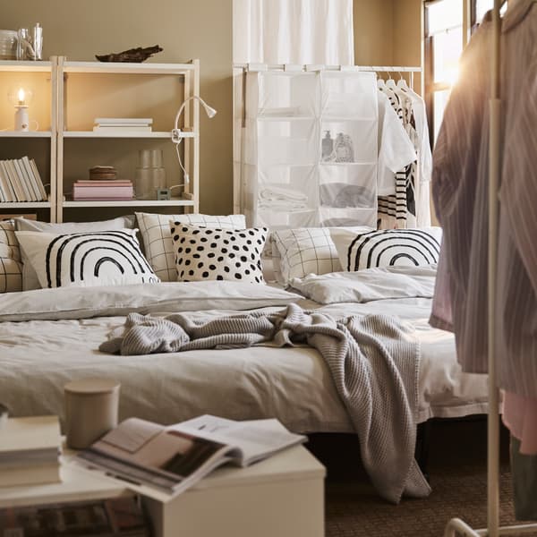 Due divani letto LYCKSELE LÖVÅS con biancheria da letto grigia e cuscini a fantasia, più un tavolino basso bianco e stand appendiabiti bianchi - IKEA.