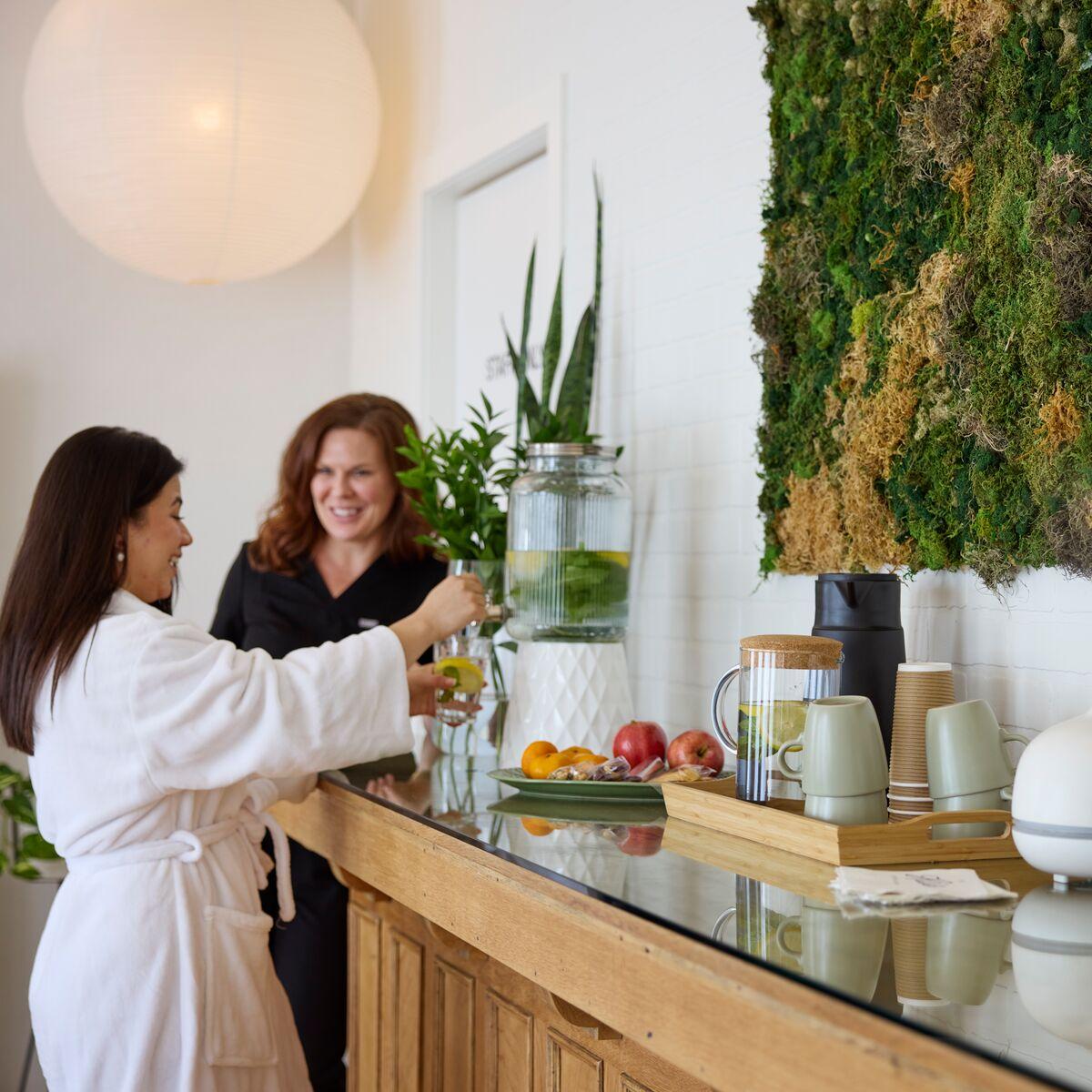 Due donne, di cui una in accappatoio che sta prendendo una bevanda da un dispenser con rubinetto VARDAGEN. L'ambiente è una sala da colazione con un mobile con sopra tazze, vassoi, frutta e altri prodotti. C'è una parete con una composizione verde.
