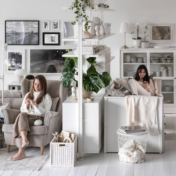 Due donne in un grande soggiorno luminoso: una è seduta su una poltrona STRANDMON beige, l'altra sul lato opposto di un elemento contenitore ELVARLI.