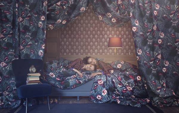 Due donne sdraiate su un letto, circondate da tessuti floreali, sullo sfondo di una carta da parati fantasia - IKEA