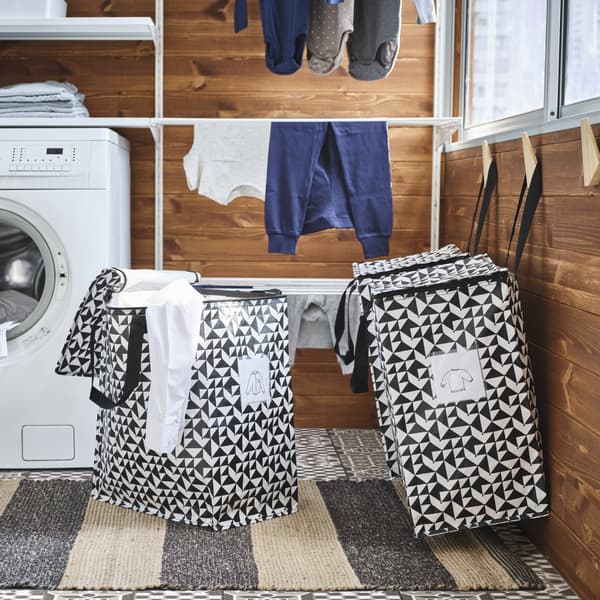Due grandi borse a motivo grafico appese a ganci di legno. Un’altra borsa, dalla quale spunta una camicia bianca, è appoggiata sul pavimento - IKEA