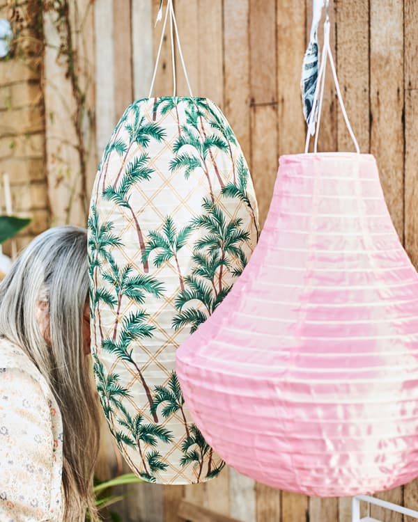 Due grandi lampade a sospensione per esterni, una rosa, l’altra con motivo con palme che nasconde parzialmente una donna con lunghi capelli grigi.