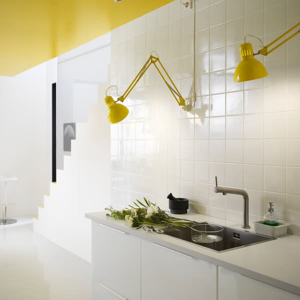 Due lampade con testa regolabile TERTIAL gialle in una cucina bianca. Sul piano, diversi fiori freschi recisi e un lavandino con dentro due contenitori di vetro. In fondo alla stanza, una scala bianca che conduce al piano superiore.