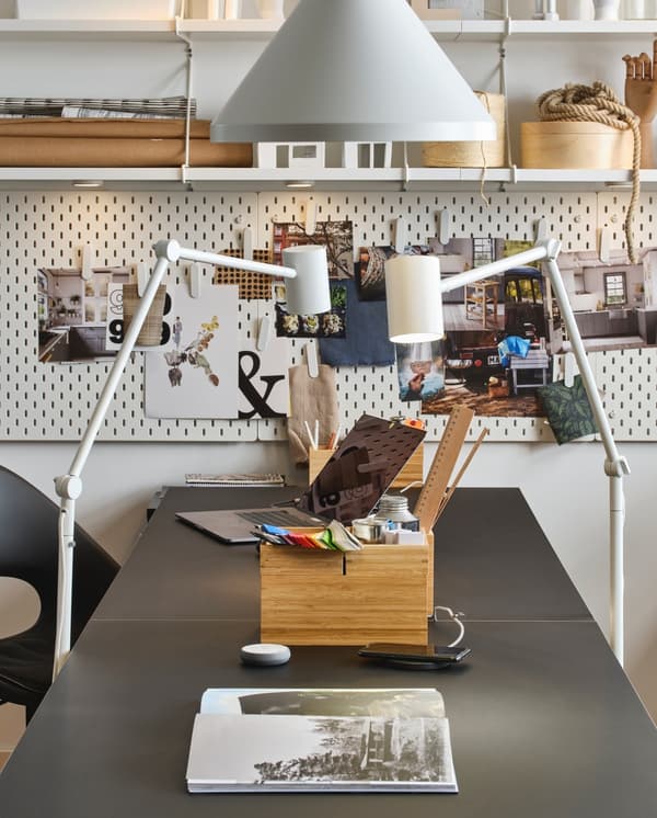 Due lampade da terra NYMÅNE bianche illuminano un tavolo scuro con un piccolo contenitore in legno pieno di accessori da ufficio, sullo sfondo  dei pannelli portaoggetti e delle mensole. 