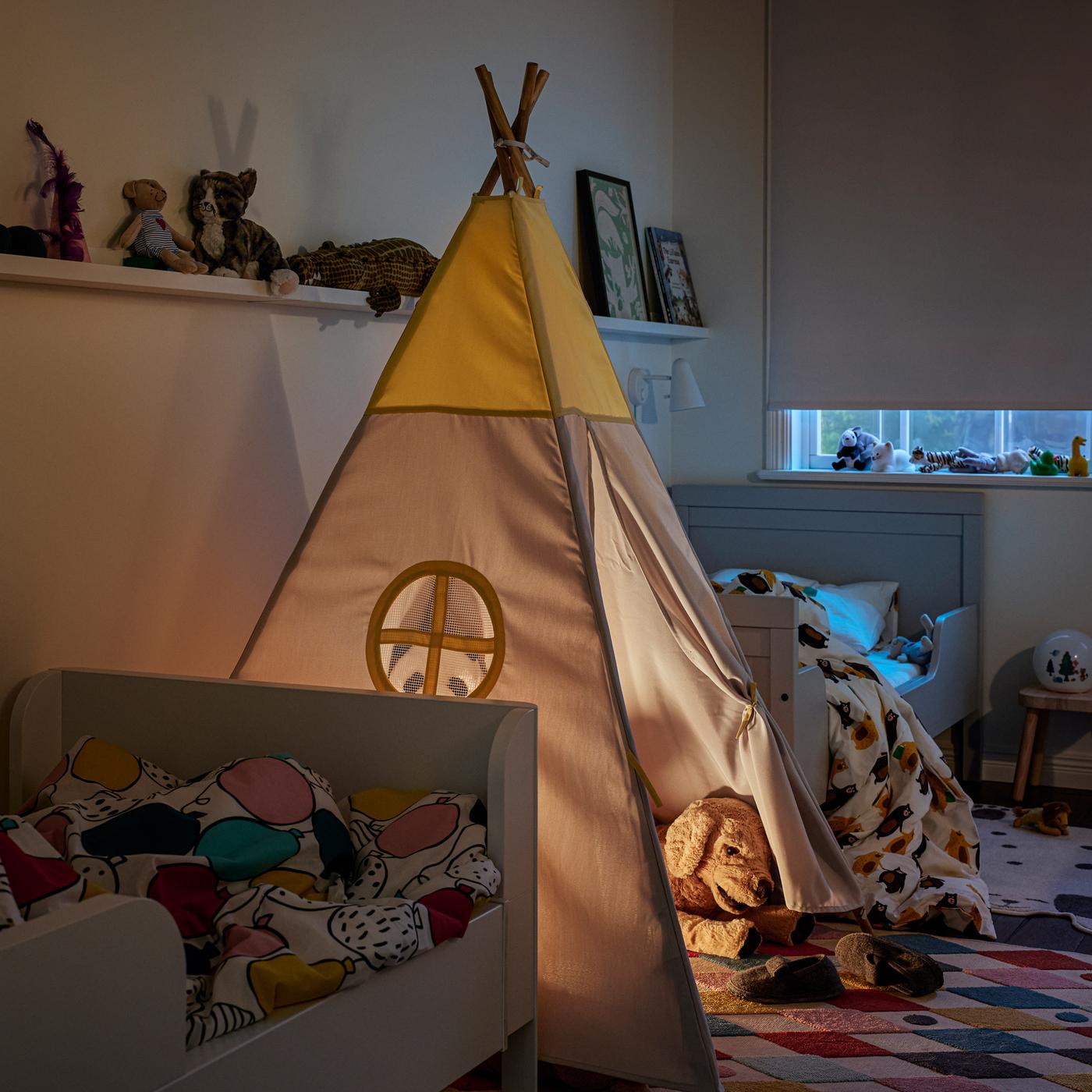 Due letti allungabili bianchi, una tenda HÖVLIG su un tappeto multicolore e una tenda a rullo bianca parzialmente abbassata.