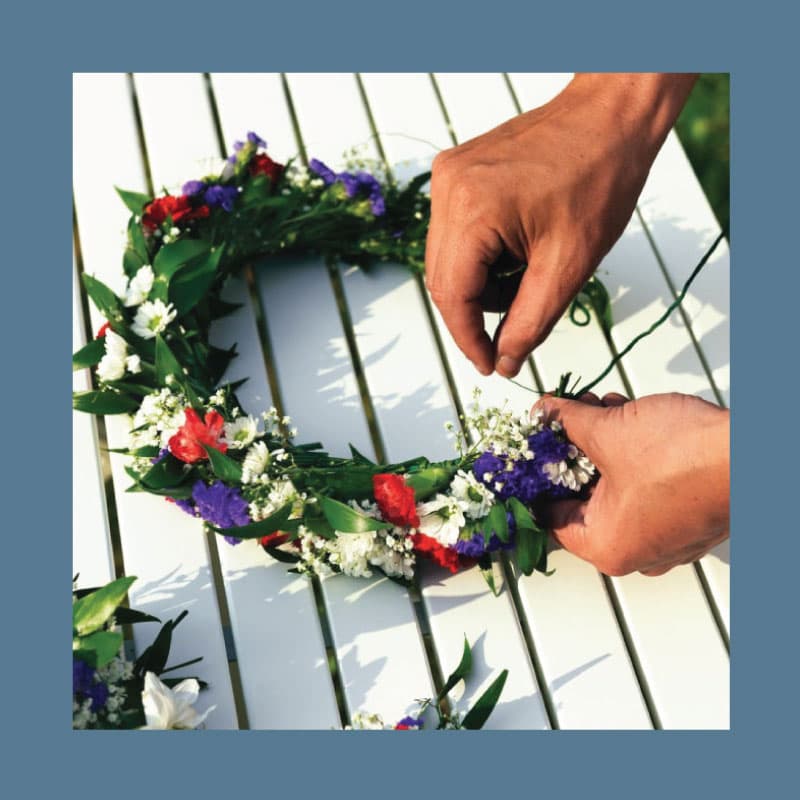 Due mani che stanno realizzando una corona di fiori sopra un tavolo bianco.
