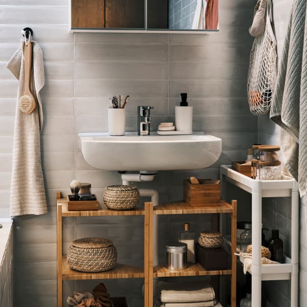 Due mobili sottolavabo/scaffali angolari in bambù sotto a un lavabo bianco, con sopra un mobile a specchio e accanto un carrello e un asciugamano.