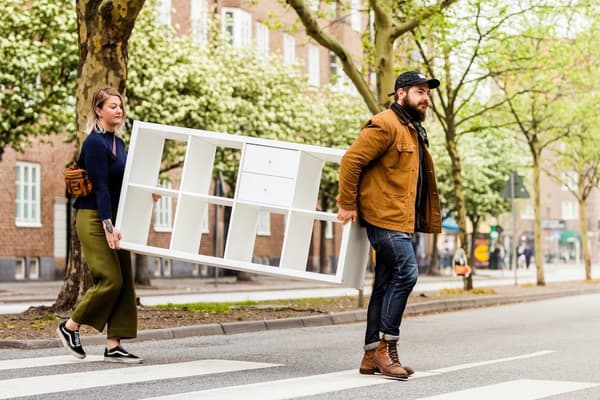 Due persone attraversano una strada trasportando uno scaffale Kallax di IKEA per rivenderlo a IKEA