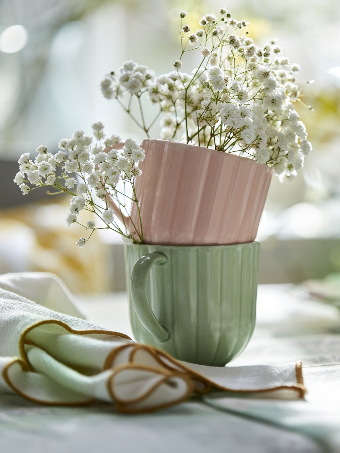 Due tazze impilate, una rosa e una verde chiaro, con un design a coste, contenenti fiori bianchi di gypsophila. Accanto, un tovagliolo verde chiaro con bordo marrone.
