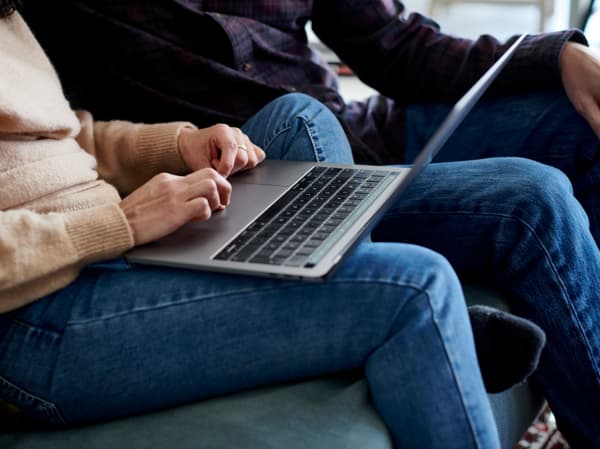 Dues persones relaxades en un sofà, una d'elles escrivint en un ordinador portàtil que descansa sobre les cames. Ambdues vesteixen texans, creant una imatge de comoditat i connexió digital.