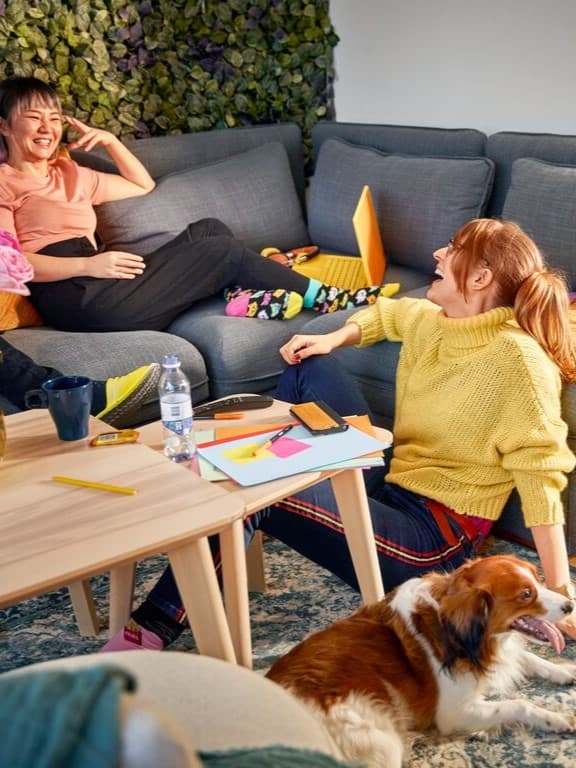 Dues persones rient i compartint un moment relaxat en un sofà, envoltades de papers i materials creatius. Un gos descansa a terra, afegint calidesa a l’escena acollidora.