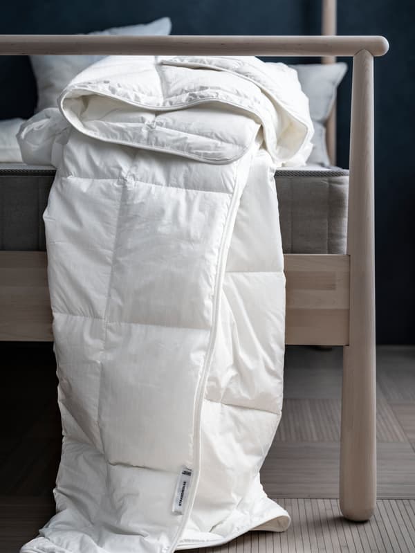 Duvet STRANDMOLKE tanpa sarung kuilt yang dilipat sembarangan terjuntai di sisi katil kayu birch GJÖRA di dalam bilik tidur bercahaya samar-samar.