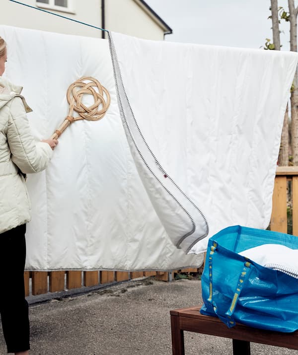 Duvet tersidai di ampaian di luar. Seorang wanita menggunakan pemukul permaidani BORSTA padanya. Di sebelahnya terdapat bangku panjang dengan beg berisi lebih banyak duvet terletak di atasnya.