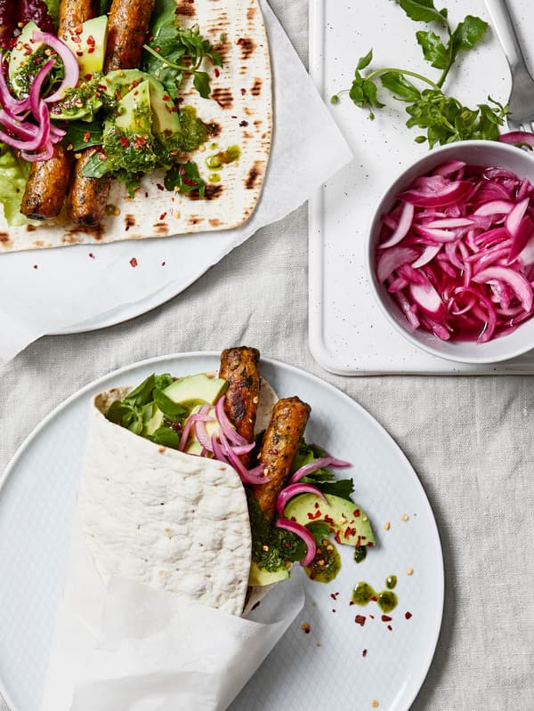 Dva bílé talíře a tenkým chlebem GRÄDDAT naplněným listovým salátem, avokádem zeleninovými hot dogya červenou cibulí, vedle na bílém prkénku je bílá miska s nakrájenou červenou cibulí.